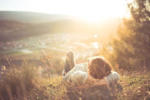 Eine Frau mit kurzen Haaren und Brille liegt rücklings im Gras auf einem Hügel im Licht der tiefstehenden Sonne. Sie hat die Arme unter dem Kopf verschränkt und schaut entspannt in den Himmel. Sie ist dabei nur von hinten zu sehen. 