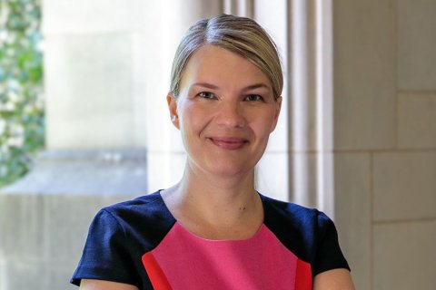 Portr&auml;t einer Frau mit kurzem, blondem, seitlich gescheiteltem Haar. Sie blickt direkt in die Kamera, die Lippen sind zu einem leichten L&auml;cheln geformt. Ihr Oberteil zeigt Farbblockierung: dunkelblaue &Auml;rmel, pink-rosafarbene Mittelpartie mit einem vertikalen roten Streifen linksseitig. Im Hintergrund ein heller Raum mit gro&szlig;em Fenster, dahinter gr&uuml;ne Pflanzen. Rechts im Bild eine beige S&auml;ule. Das Licht ist weich und nat&uuml;rlich, die Atmosph&auml;re ruhig.