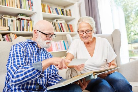 Ein &auml;ltere Dame zeigt einem &auml;lteren Herrn Fotos in einem Fotoalbum. Sie sitzen auf Sesseln vor einem B&uuml;cherregal. Beide tragen Brillen. Die Same l&auml;chelt, w&auml;hrend der Herr etwas unsicher mit einem Finger auf ein Foto zeigt. 