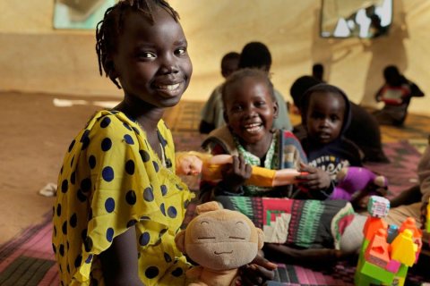 Ein M&auml;dchen mit gelber Bluse und dunklen Punkten sitzt auf einer Matte und l&auml;chelt in die Kamera. In ihrer Hand h&auml;lt sie ein beigefarbenes Pl&uuml;schtier mit Affengesicht. Neben ihr sitzt ein j&uuml;ngeres lachendes M&auml;dchen mit einer Puppe in der Hand. Weitere Kinder spielen im Hintergrund in einem hellen Zelt. Die Atmosph&auml;re wirkt lebendig und freundlich.
