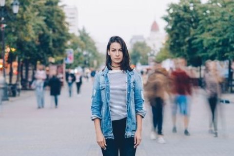 Eine junge Frau mit langen dunklen Haaren steht auf einer belebten Straße. Sie trägt eine gestreifte Bluse, eine offene Jeansjacke und schwarze Hosen. Um sie herum gehen viele Menschen vorbei, deren Bewegungen verschwommen wirken, während sie selbst scharf und ruhig im Zentrum des Bildes bleibt. Im Hintergrund sind Bäume, Häuser und Lichter der Stadt zu erkennen.