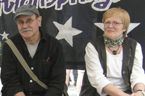 Zwei &auml;ltere Personen sitzen nebeneinander auf einer Bank vor einem schwarzen Banner mit wei&szlig;en Sternen und dem Schriftzug &bdquo;&hellip;tionspflege". Links ein Mann mit grauer Schirmm&uuml;tze, Brille, grauem Schnurrbart, schwarzem Hemd und Umh&auml;ngetasche. Rechts eine Frau mit kurzem blonden Haar, Brille, wei&szlig;em Pullover, dunkler Weste und gemustertem Halstuch. Beide blicken leicht l&auml;chelnd nach vorn.