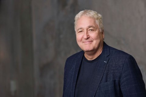 Portr&auml;taufnahme eines Mannes, etwa sechzig Jahre alt, im Brustbereich angeschnitten. Er steht leicht nach links gedreht, den Kopf zur Kamera gewandt. Sein Haar ist wei&szlig; und wellig nach hinten gek&auml;mmt. Er l&auml;chelt dezent, die Augen sind leicht zusammengekniffen. Er tr&auml;gt ein dunkelblaues Sakko mit hellem Karomuster &uuml;ber einem dunkelblauen Rundhals-T-Shirt. Der Hintergrund zeigt eine unscharfe graue Fl&auml;che mit beigen Schlieren, vermutlich Beton. Weiches Licht von links.