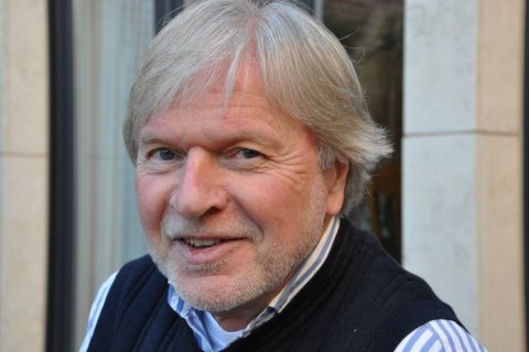 Ein &auml;lterer Mann mit grauen Haaren und kurzem Bart blickt l&auml;chelnd in die Kamera. Er tr&auml;gt ein dunkelblaues Oberteil &uuml;ber einem hellblauen Hemd mit Streifen. Im Hintergrund sind unscharf helle Steinw&auml;nde und ein Fenster zu erkennen. Das Bild ist eine Nahaufnahme, der Fokus liegt auf dem Gesicht des Mannes, das freundlich und offen wirkt.