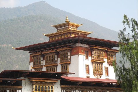 Ein traditionelles Klostergeb&auml;ude in Bhutan mit wei&szlig;en Mauern, reich verzierten gold-roten Holzelementen und mehreren gestaffelten D&auml;chern. Auf der Spitze thront ein vergoldeter Turm. Im Hintergrund ragen bewaldete Berge empor, w&auml;hrend ein Baumzweig rechts ins Bild ragt.