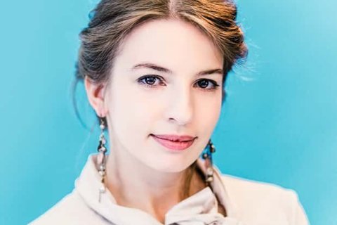 Halbnahes Porträt einer jungen Frau vor blauem Hintergrund. Sie trägt eine helle Bluse mit Schleife, dezenten Lippenstift und lange Ohrringe und blickt ruhig und freundlich in die Kamera.