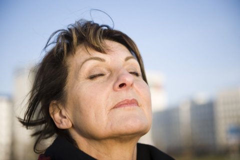 Im Vordergrund ist der Kopf einer Frau vor blauem Himmel und unscharf gehaltenen Geb&auml;uden im Hintergrund zu sehen. Die Frau scheint den Kopf in den Wind zu halten. Die Haare sind ein wenig zerzaust, sie hat Augen und Mund geschlossen und scheint durch die Nase tief durchzuatmen. 