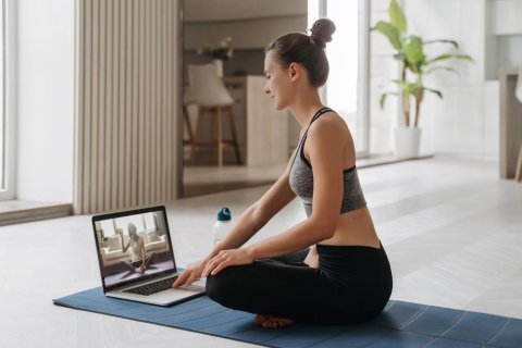 Eine Frau hockt im Schneidersitz auf einer Yogamatte. Vor sich hat sie ein Laptop stehen, in dessen Bildschirm eine andere Frau im Schneidersitz zu sehen ist, die in die Kamera winkt. 