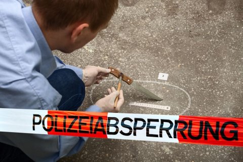 Ein Mann mit Handschuhen bearbeitet hinter eine Polizeiabsperrung kniend ein Messer mit einem Pinsel, welches am Boden in einem Kreidekreis gelegen hatte. 