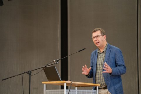 Ein mittelgro&szlig;er Mann mittleren Alters steht an einem schmalen Redepult. Vor ihm ein aufgeklappter Laptop, davor ein langes Mikrofon auf Stativ. Er tr&auml;gt Brille, blaues Sakko, kariertes Hemd und beige Hose. Seine H&auml;nde sind vor dem K&ouml;rper ausgebreitet, als ob er etwas betont erkl&auml;rt. Der Hintergrund ist eine einfarbige, graue Saalwand.