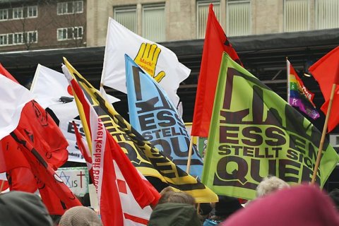 In einer Menschenmenge sind viele Fahnen dicht beieinander zu sehen. Dominant ist eine gro&szlig;e gr&uuml;ne Fahne mit der Aufschrift &bdquo;Essen stellt sich quer&ldquo;. Daneben ragen weitere Banner in Rot, Wei&szlig;, Gelb und Blau empor. Die K&ouml;pfe der Demonstrierenden sind nur teilweise im unteren Bildrand erkennbar. Im Hintergrund steht eine graue Geb&auml;udefassade mit Fenstern.