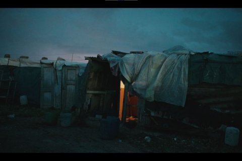 In der D&auml;mmerung stehen einfache H&uuml;tten aus Holz, Planen und Plastikfolien auf einem dunklen, schlammigen Gel&auml;nde. Eine T&uuml;r ist ge&ouml;ffnet, warmes Licht f&auml;llt aus dem Inneren. Vor den Unterk&uuml;nften stehen Kanister und Beh&auml;lter. Der Himmel ist blaugrau, die Szene wirkt kalt, provisorisch und verlassen.