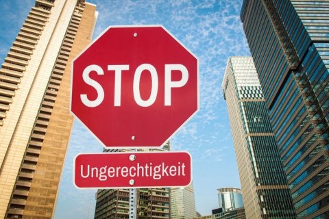 Rotes achteckiges Stoppschild mit wei&szlig;er Aufschrift &bdquo;STOP&ldquo;, darunter ein kleineres rotes Zusatzschild mit dem Wort &bdquo;Ungerechtigkeit&ldquo;. Die Schilder stehen aus der Froschperspektive fotografiert zwischen Hochh&auml;usern. Links eine sandfarbene Fassade mit Balkonreihen, rechts blaugr&uuml;n spiegelnde Glast&uuml;rme. Dar&uuml;ber blauer Himmel mit feinen Schleierwolken. Das Rot der Schilder hebt sich deutlich von der k&uuml;hlen Architektur ab.
