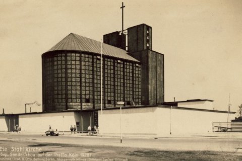 Historische Schwarz-Wei&szlig;-Aufnahme eines modernen Kirchenbaus. Der Bauk&ouml;rper besteht aus einem hohen, runden Glasturm mit kegelf&ouml;rmigem Dach und engem Raster dunkler Fenstersprossen. Rechts erhebt sich ein kantiger, dunkler Doppelturm, auf dessen Spitze ein Kreuz steht; darunter h&auml;ngt eine Glocke. Der helle Sockel ist flach gedeckt. Davor kleine Personengruppen, ein offener Wagen, ein Fahnenmast. Schriftzug: &bdquo;Die Stahlkirche (Evgl. Sonderschau auf der Pressa K&ouml;ln 1928)". Heller, dunstiger Himmel.