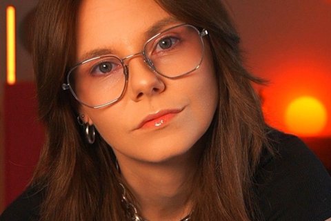 Portr&auml;t der Autorin Anja Heyelmann in Nahaufnahme. Sie blickt direkt in die Kamera, der Kopf ist leicht nach vorn geneigt. Sie tr&auml;gt eine gro&szlig;e Brille mit d&uuml;nnem silbernem Metallgestell, schulterlanges hellbraunes Haar mit Pony, einen silbernen Ohrring und eine grobgliedrige Silberkette zum schwarzen Oberteil. Der Mund ist geschlossen, ein Mundwinkel leicht angehoben. Der Hintergrund ist unscharf und in warmes Rot und Orange getaucht, rechts leuchtet ein runder gelber Lichtpunkt.
