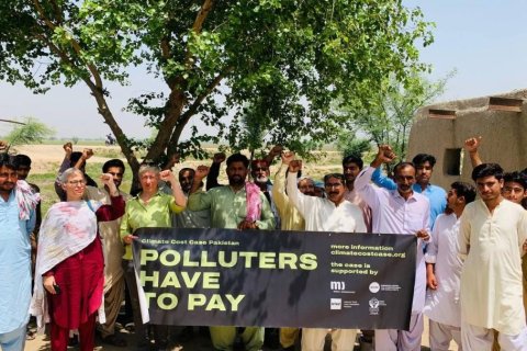 Eine Gruppe M&auml;nner und zwei Frauen stehen unter einem Baum vor einer Lehmmauer und recken die F&auml;uste in die H&ouml;he. Vorn halten sie ein schwarzes Banner mit der Aufschrift &bdquo;Climate Cost Case Pakistan &ndash; Polluters have to pay&ldquo; und dem Hinweis &bdquo;more information: climatecostcase.org&ldquo;. Viele tragen helle oder farbige Shalwar Kamiz, einige T&uuml;cher &uuml;ber der Schulter. Links &ouml;ffnet sich eine flache, sonnenhelle Ebene, das Licht f&auml;llt glei&szlig;end durch das Laub.